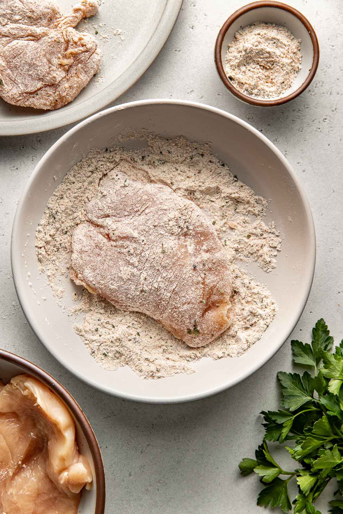 Overhead view of a chicken breast being dredged in a flour and seasoning mixture. 