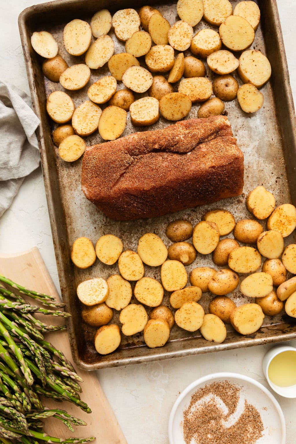 Overhead view of a sheet pan with a seasoned pork tenderloin and baby potatoes on it, ready for baking. 