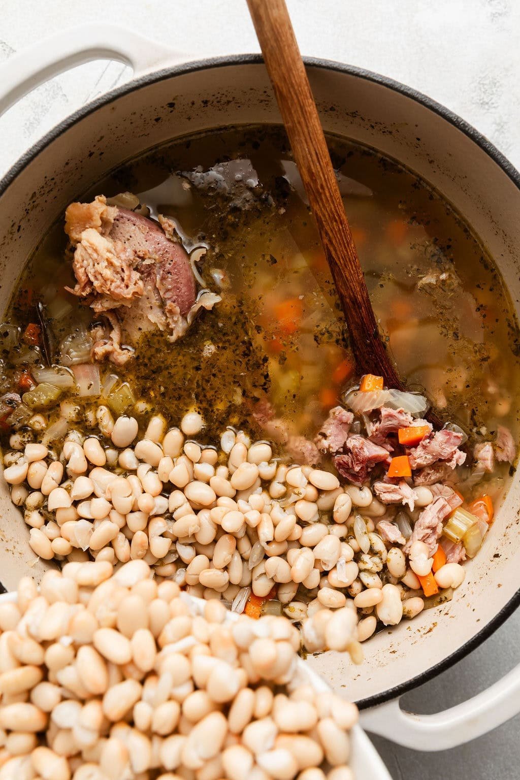 Overhead view of ingredients for Ham and Bean Soup going into a Dutch oven with a wooden spoon in it. 