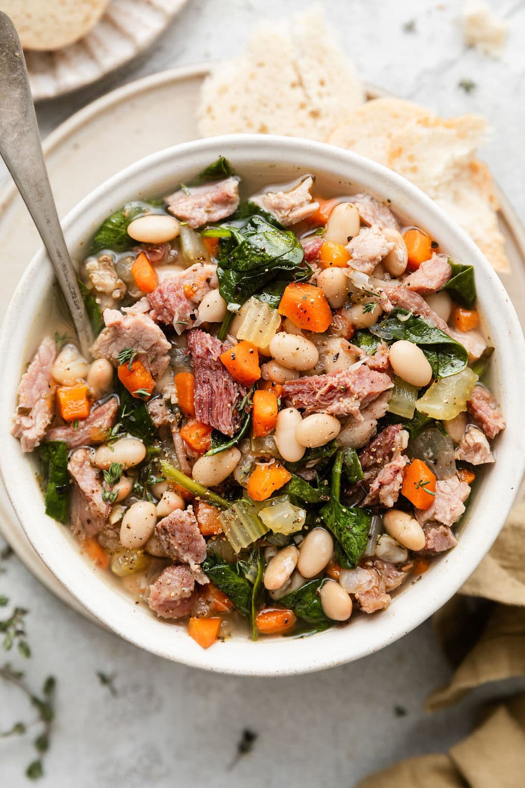 Overhead view of a white bowl filled with Ham And Bean Soup and pieces of bread on the side.