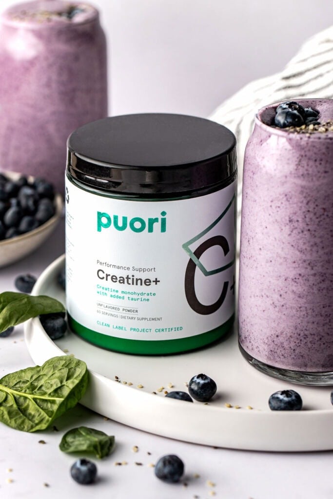 A canister of creatine next to a deep purple smoothie surrounded by spinach leaves and blueberries. 