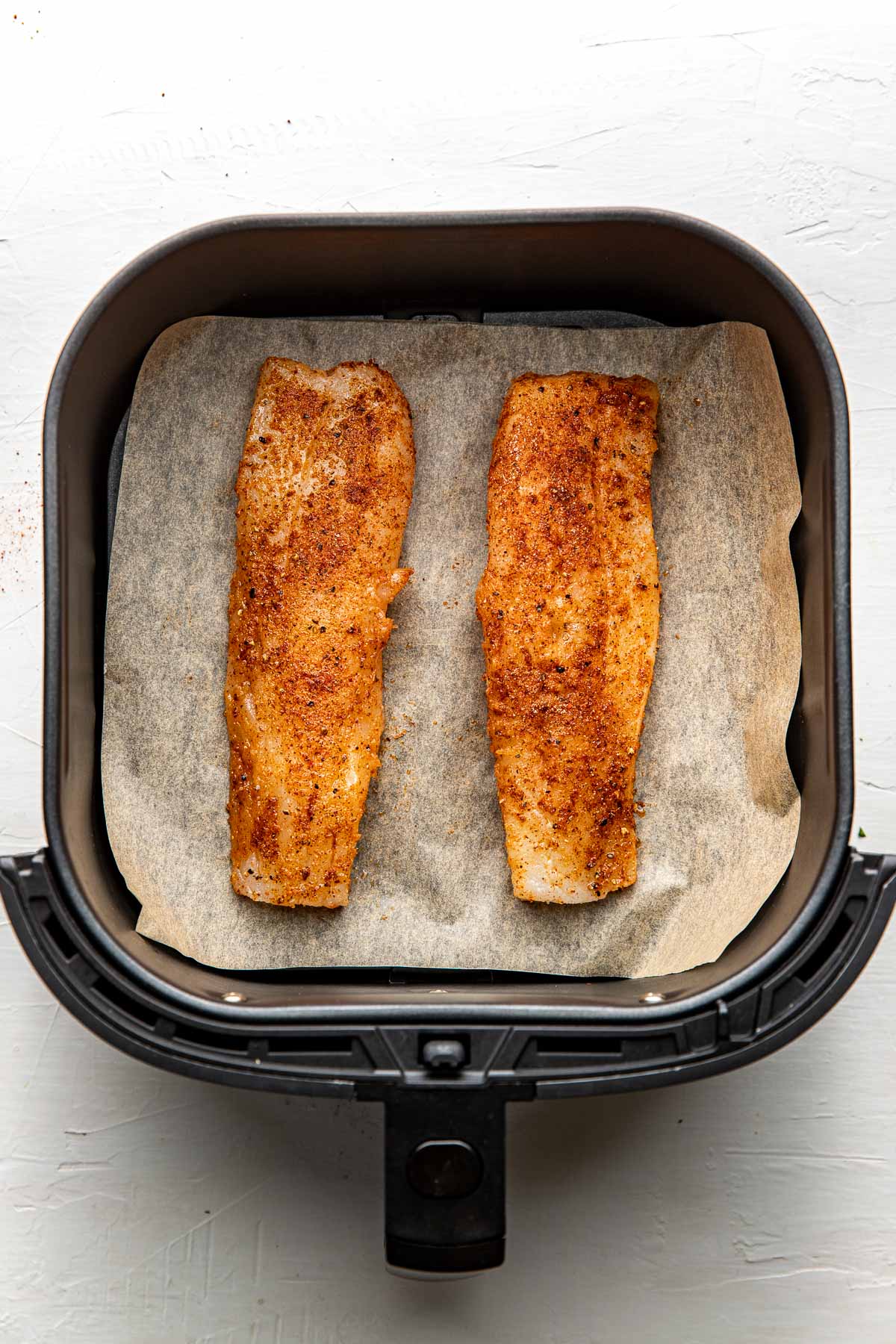 Overhead view of seasoned cod fillets in an air fryer basket lined with parchment paper ready for baking.