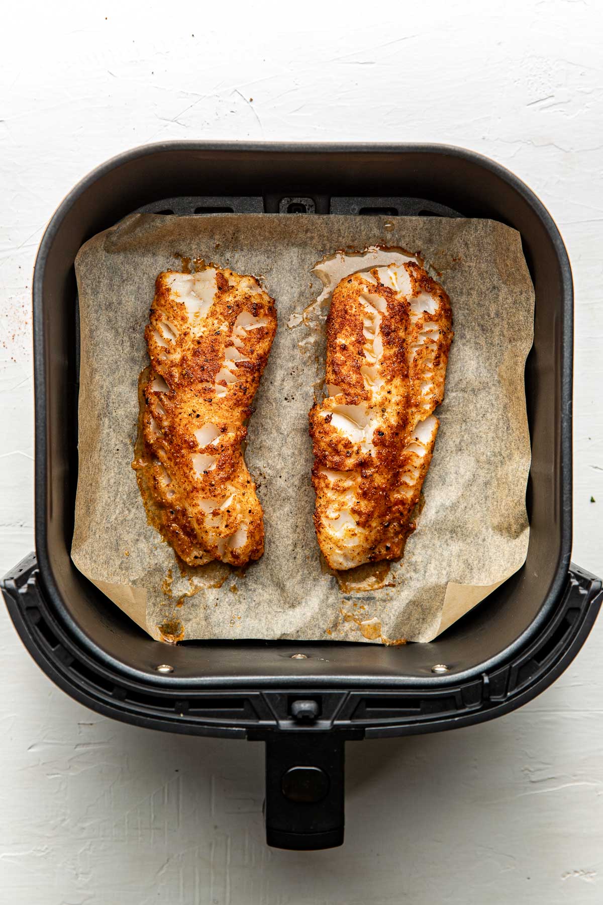 Overhead view of freshly baked seasoned cod on parchment paper in an air fryer basket.