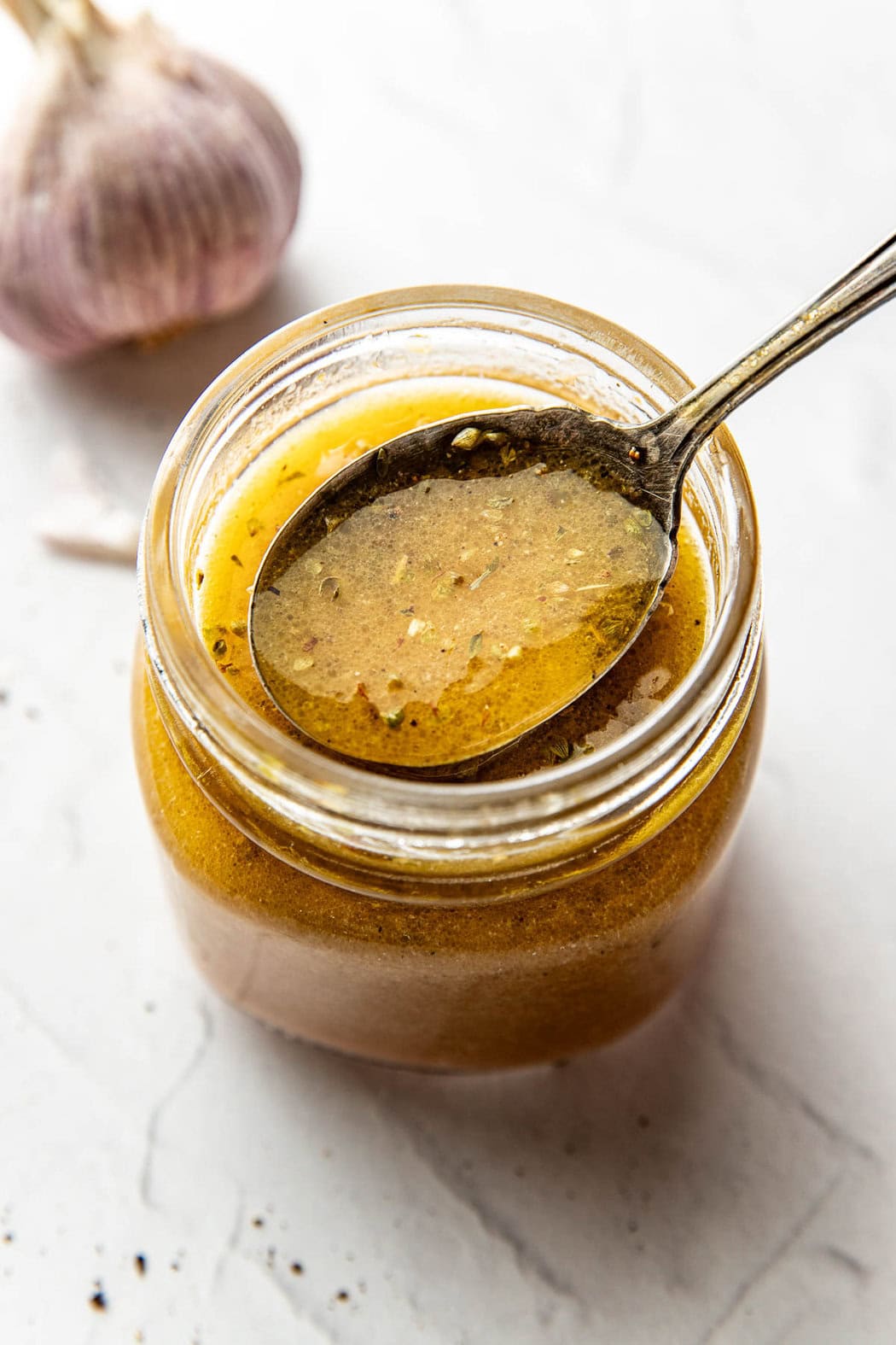 Overhead view of a glass jar of Red Wine Vinaigrette with a spoon bringing up a scoop to show the rich color and texture of the herbs. 