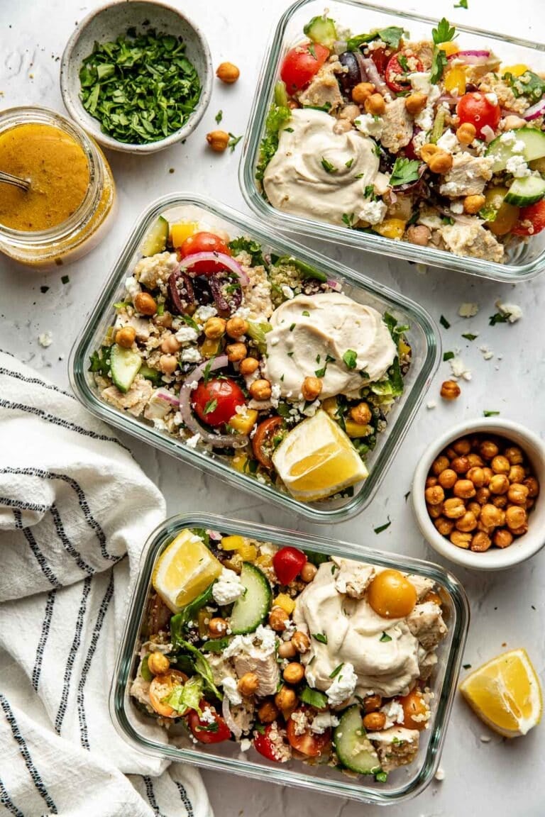 Overhead view of three glass meal prep containers filled with mediterranean chopped salad topped with hummus and feta crumbles.