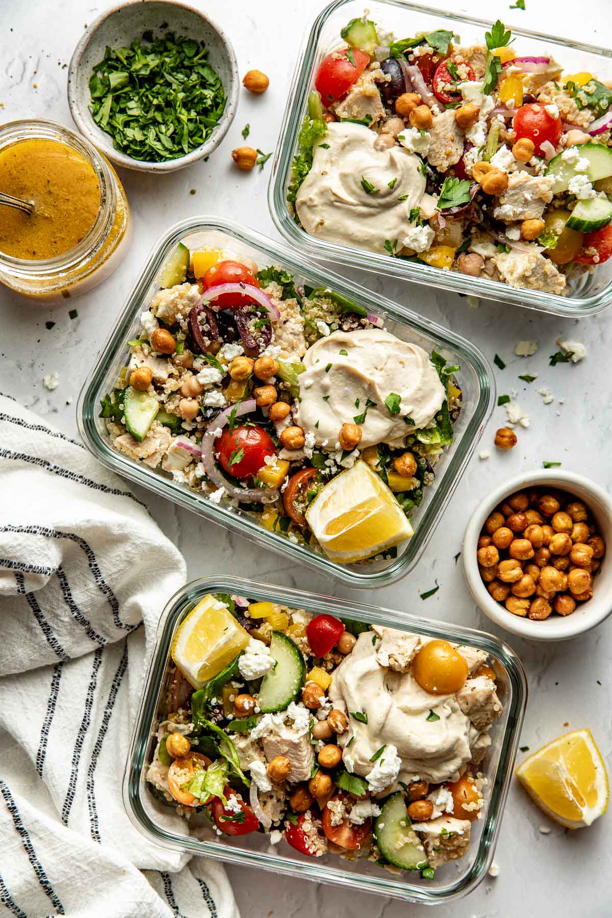 Overhead view of three glass meal prep containers filled with Chopped Mediterranean Salad with Chicken and a dollop of hummus in each container. 