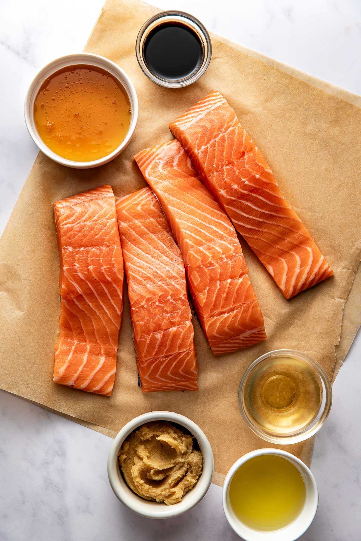 Overhead view of 4 salmon fillets on parchment paper surrounded by liquid ingredients in small dishes. 
