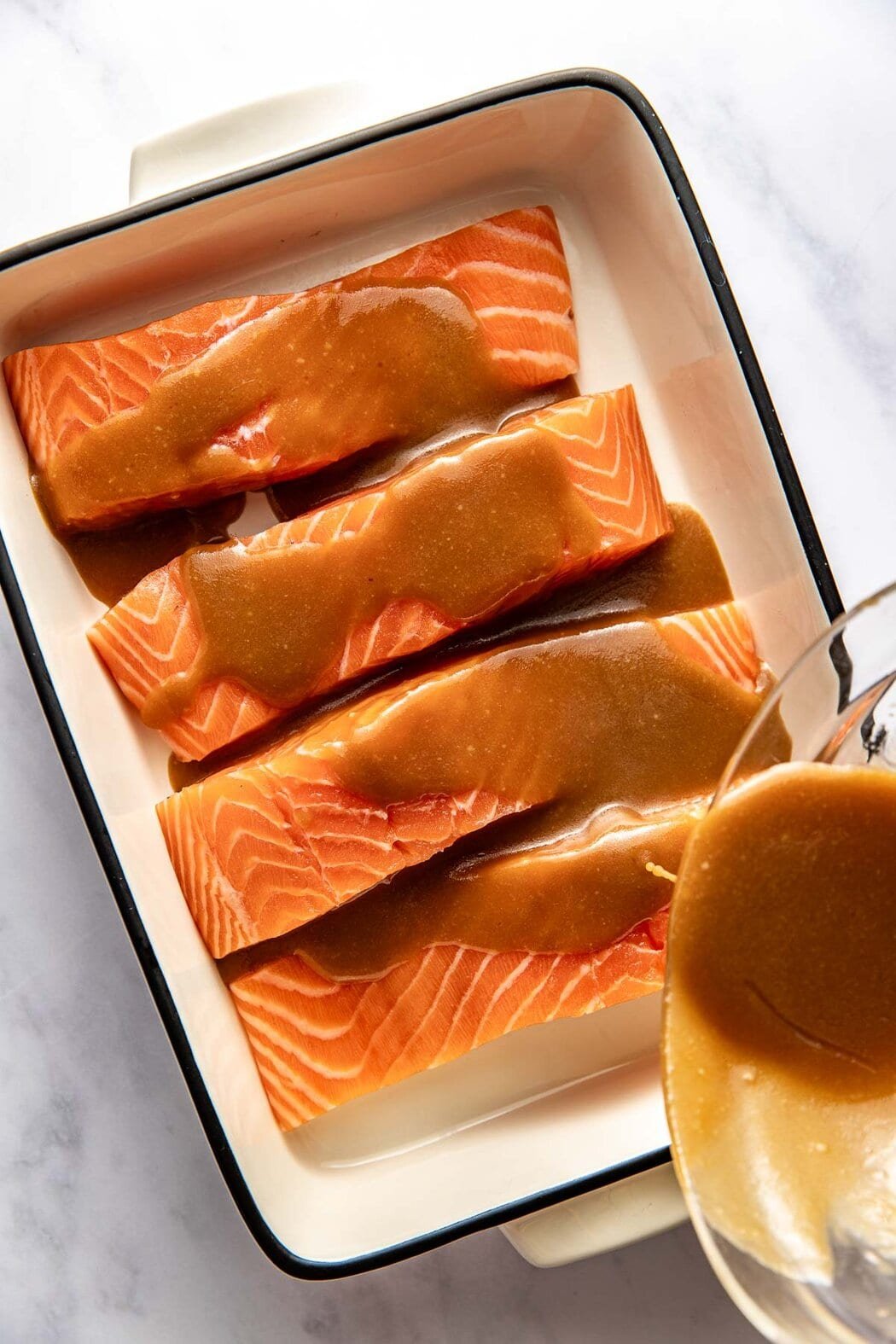 Overhead view of 4 salmon fillets in a pan with Miso sauce being poured over the top. 