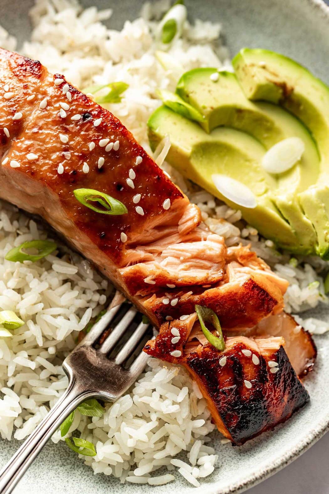 Close up view of a fork in a piece of Miso-Glazed Salmon on top of rice topped with green onions. 