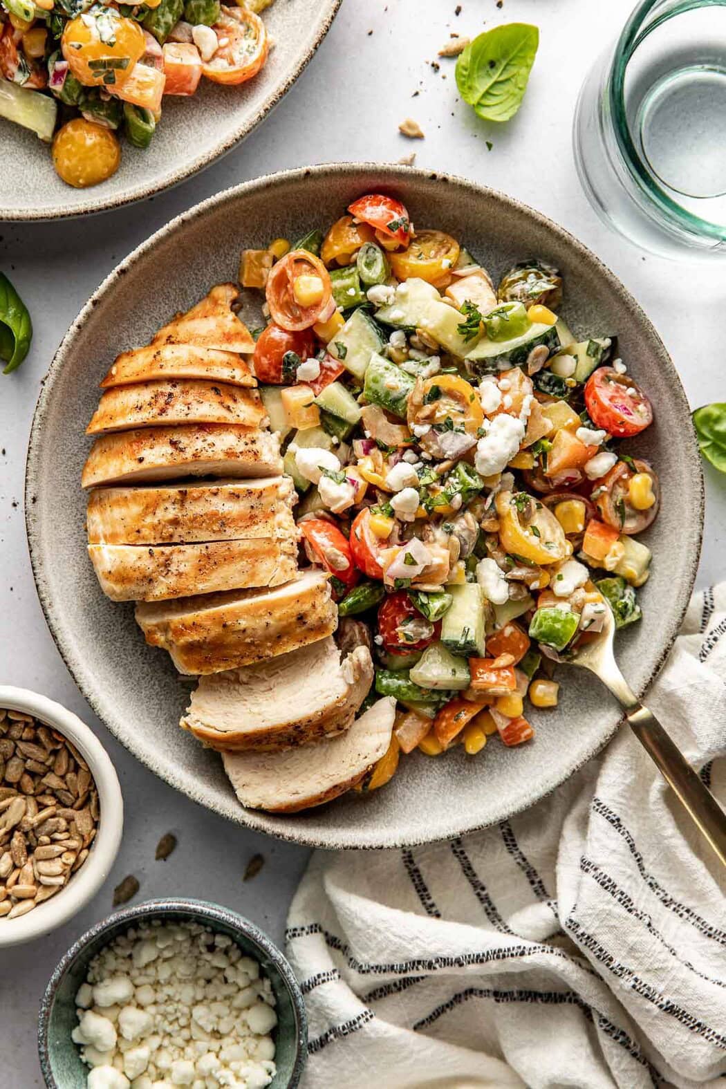 Overhead view of a plate of colorful summer salad topped with creamy goat cheese next to a sliced grilled chicken breast