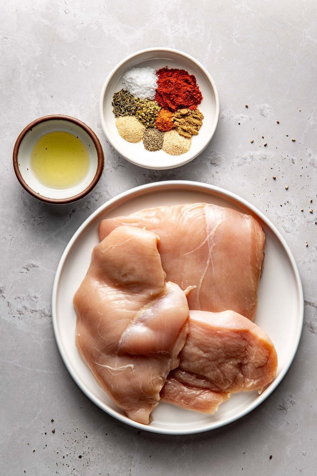 Overhead view of a small dish of oil, a small dish of seasonings, and a plate with three chicken breasts on it.