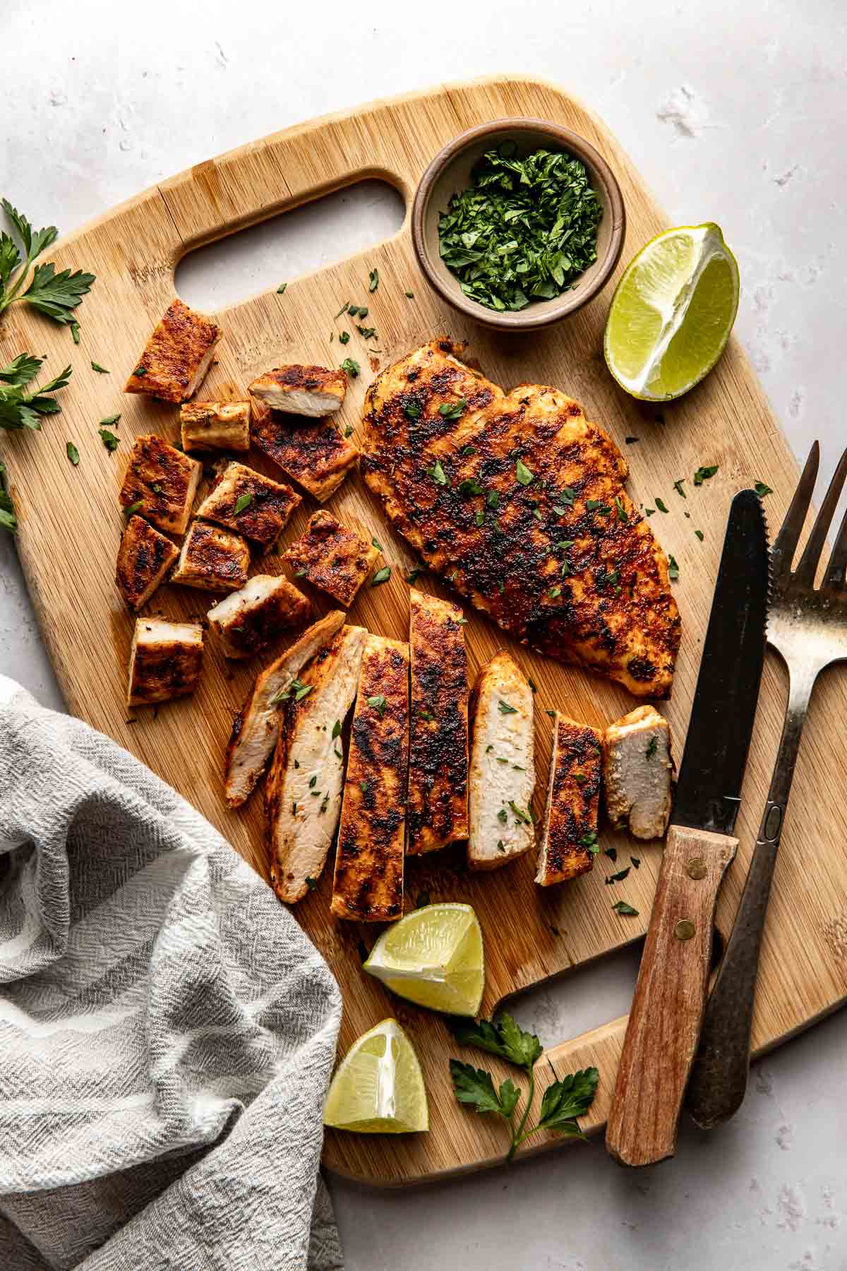 Overhead view of a wooden cutting board filled with Blackened Chicken breasts and pieces cut, next to a fork and knife and limes surrounding the chicken.