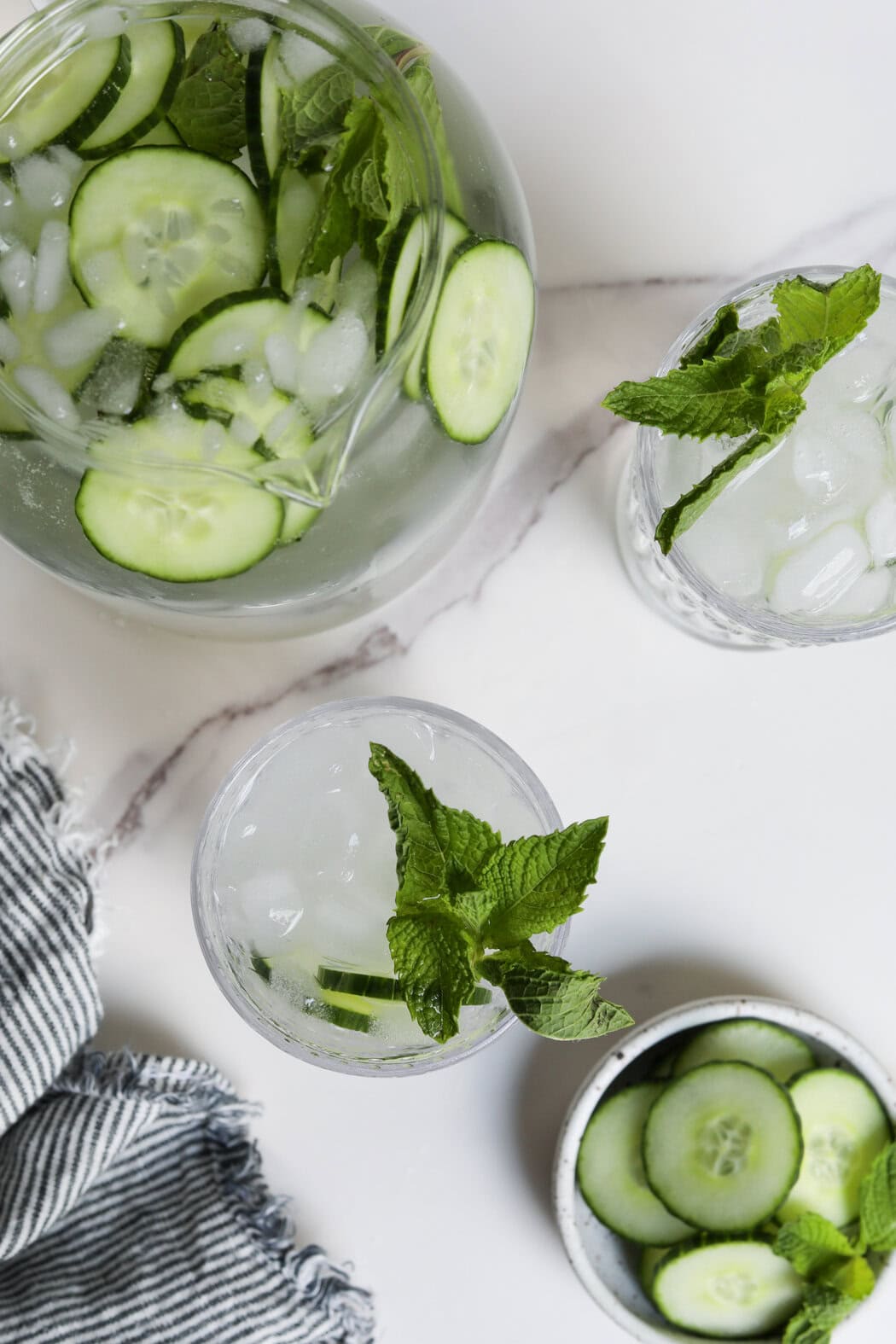 Overhead view of a water pitcher and two glasses filled with cucumber water and topped with ice, sliced cucumbers, and fresh mint leaves. 