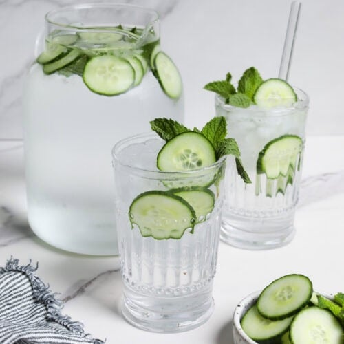 Close up view of a water pitcher and two glasses filled with fresh sliced cucumbers and mint leaves