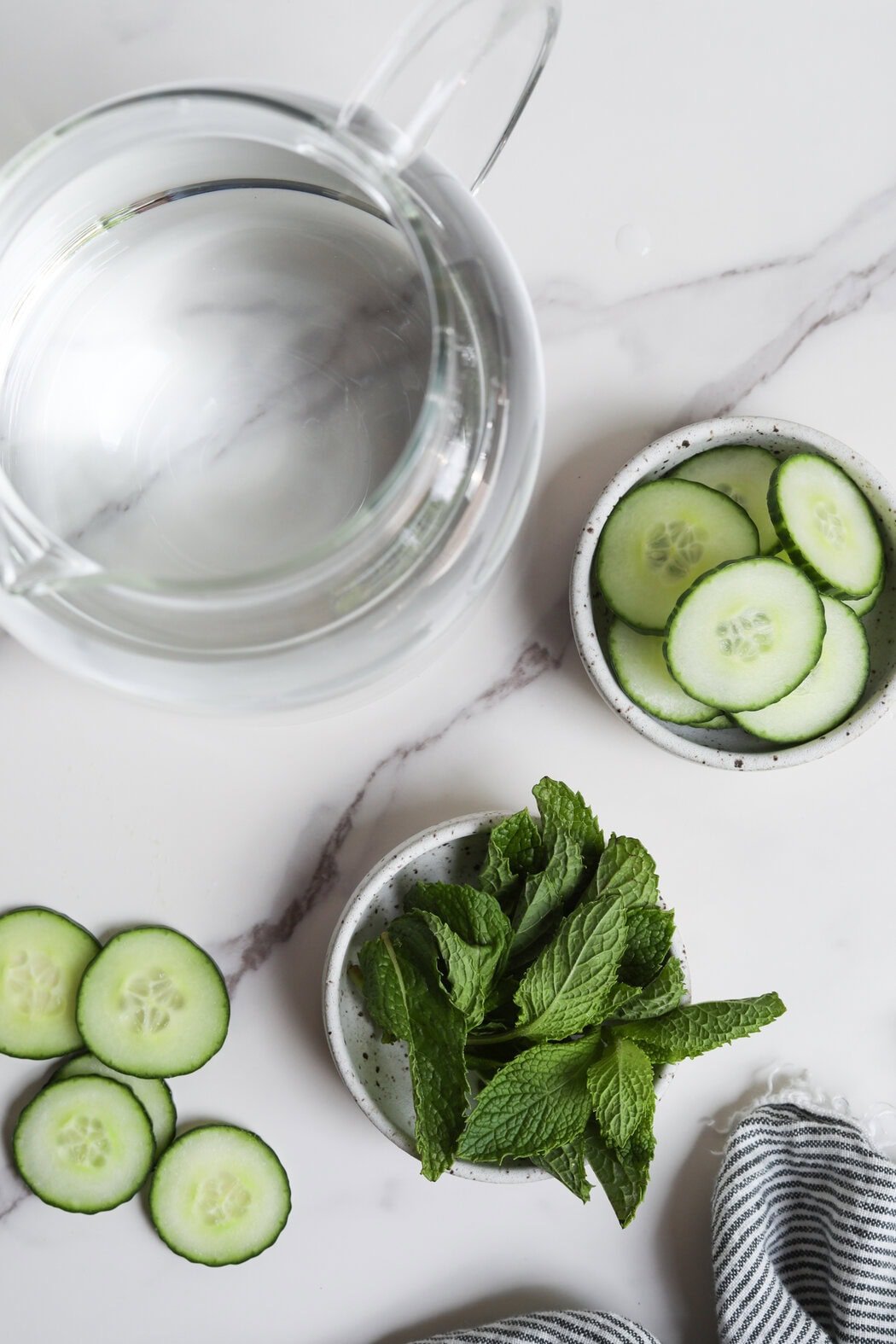 Overhead view of a glass water pitcher filled with water and two small dishes filled with sliced cucumbers and fresh mint leaves. 