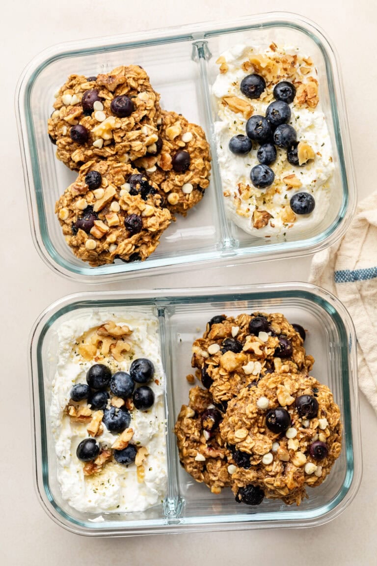 Overhead view of two glass meal prep containers filled with blueberry oatmeal breakfast cookies and cottage cheese topped with granola and blueberries.