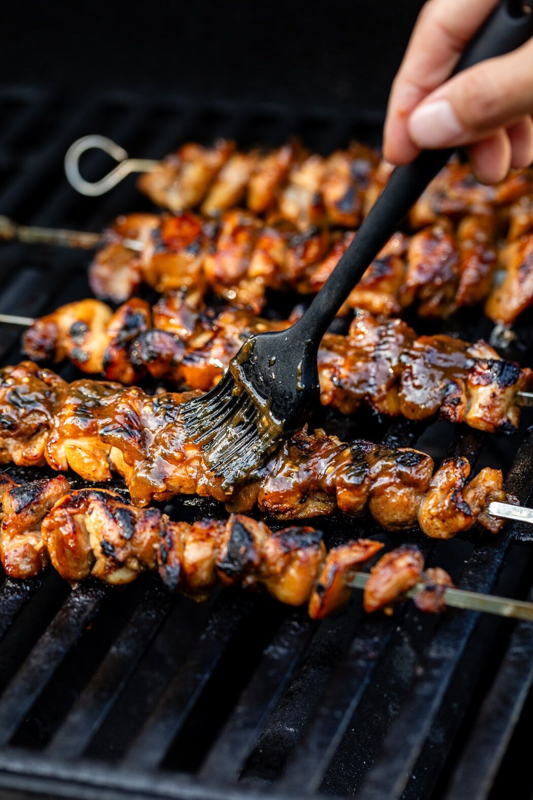 Close up view of a hand brushing Korean BBQ sauce over chicken skewers on a grill 