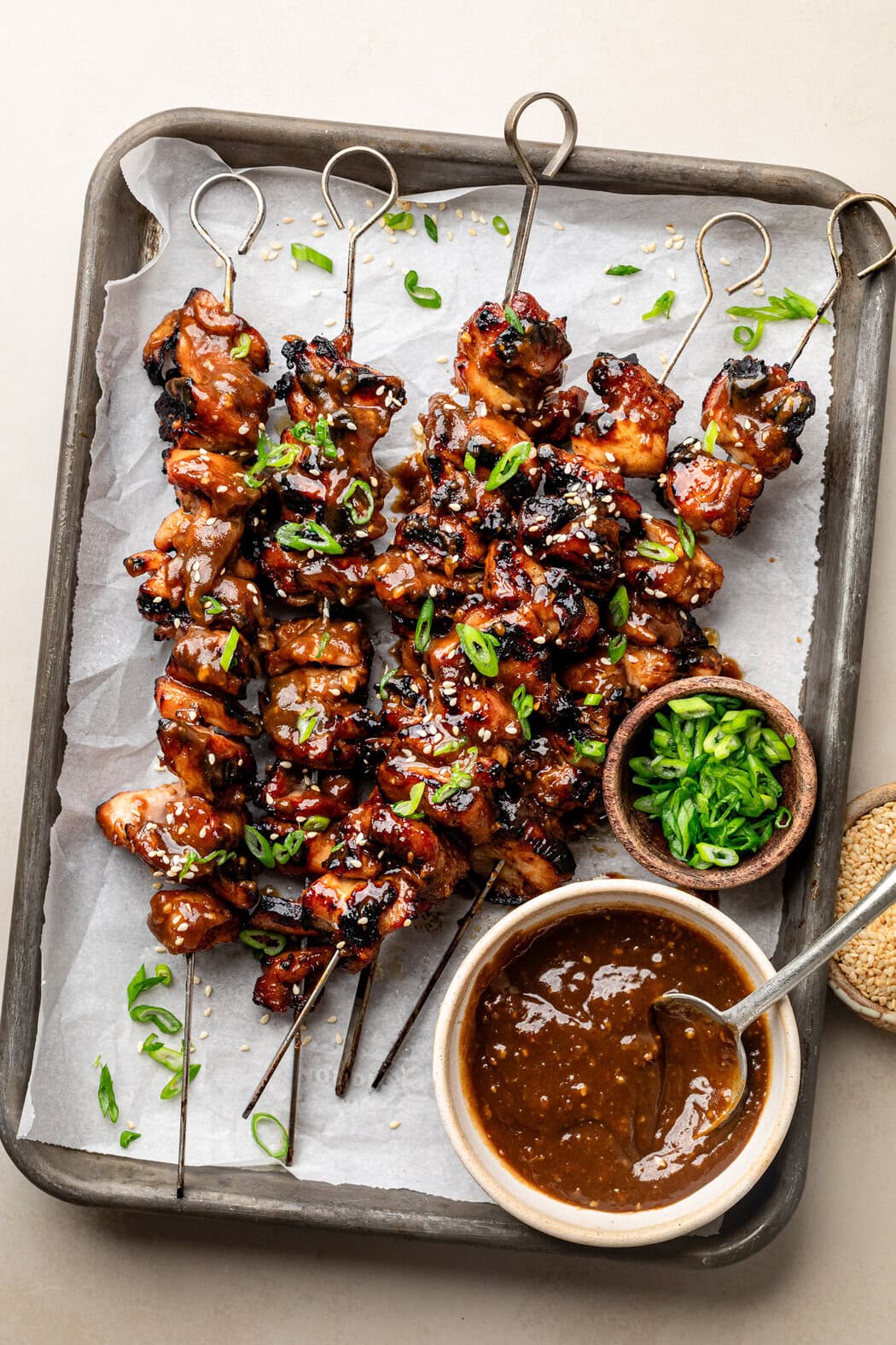 Overhead view of a sheet pan lined with white parchment paper and topped with metal skewers of Grilled Korean BBQ Chicken topped with green onions and sesame seeds.