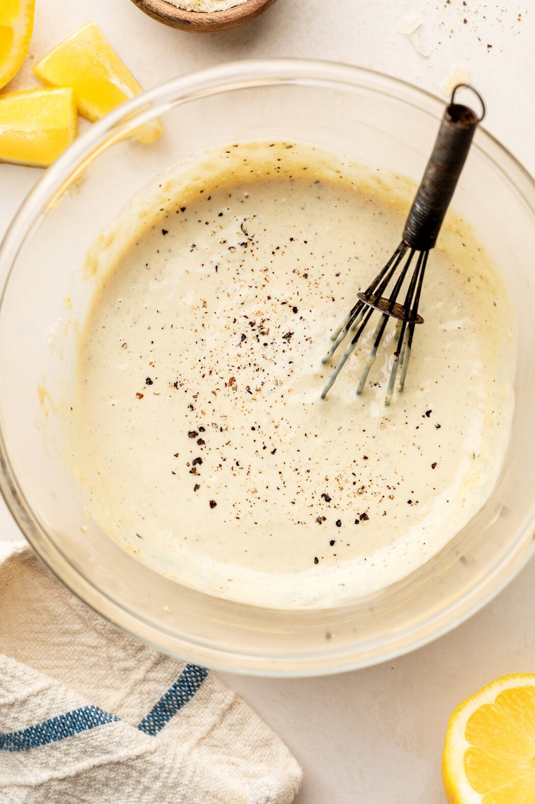Overhead view of a glass bowl of homemade Caesar dressing topped with black pepper and a whisk on the side of the bowl. 