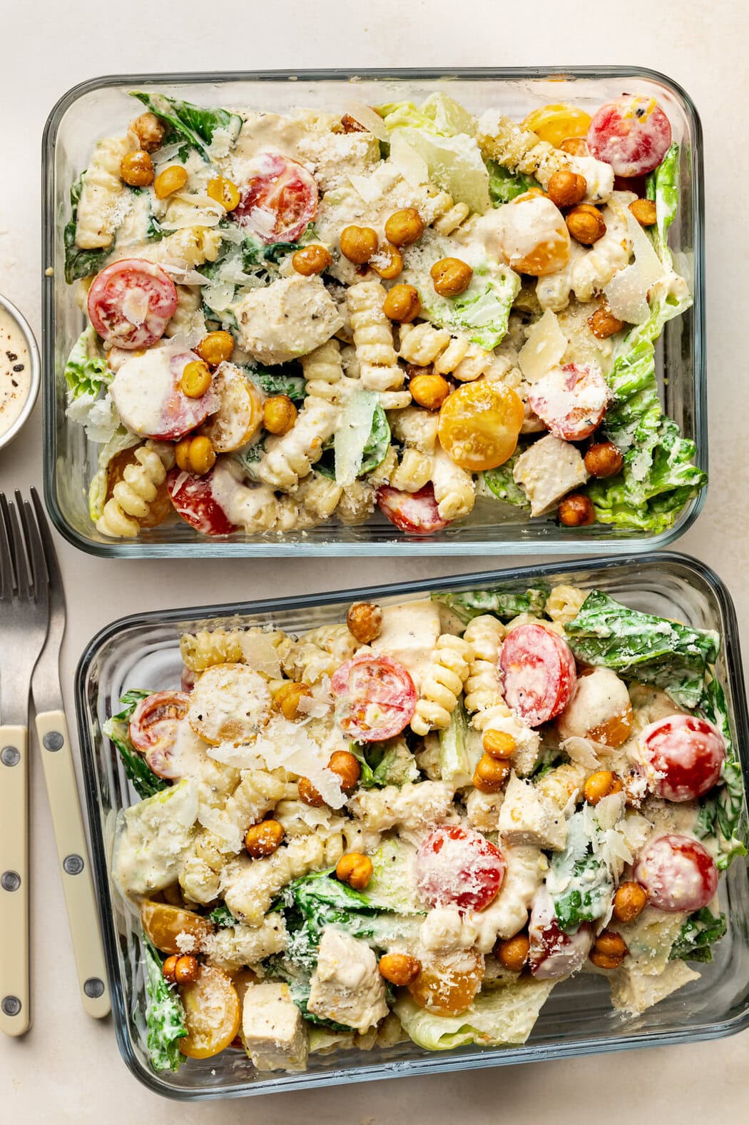 Overhead view of two glass meal prep containers filled with Chicken Caesar Pasta Salad topped with grated parmesan cheese and crisp roasted chickpeas. 