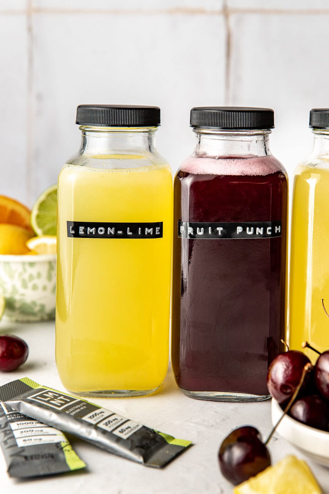 Profile view of two glass bottles of Homemade Gatorade labeled lemon lime and fruit punch surrounded by fresh fruit. 