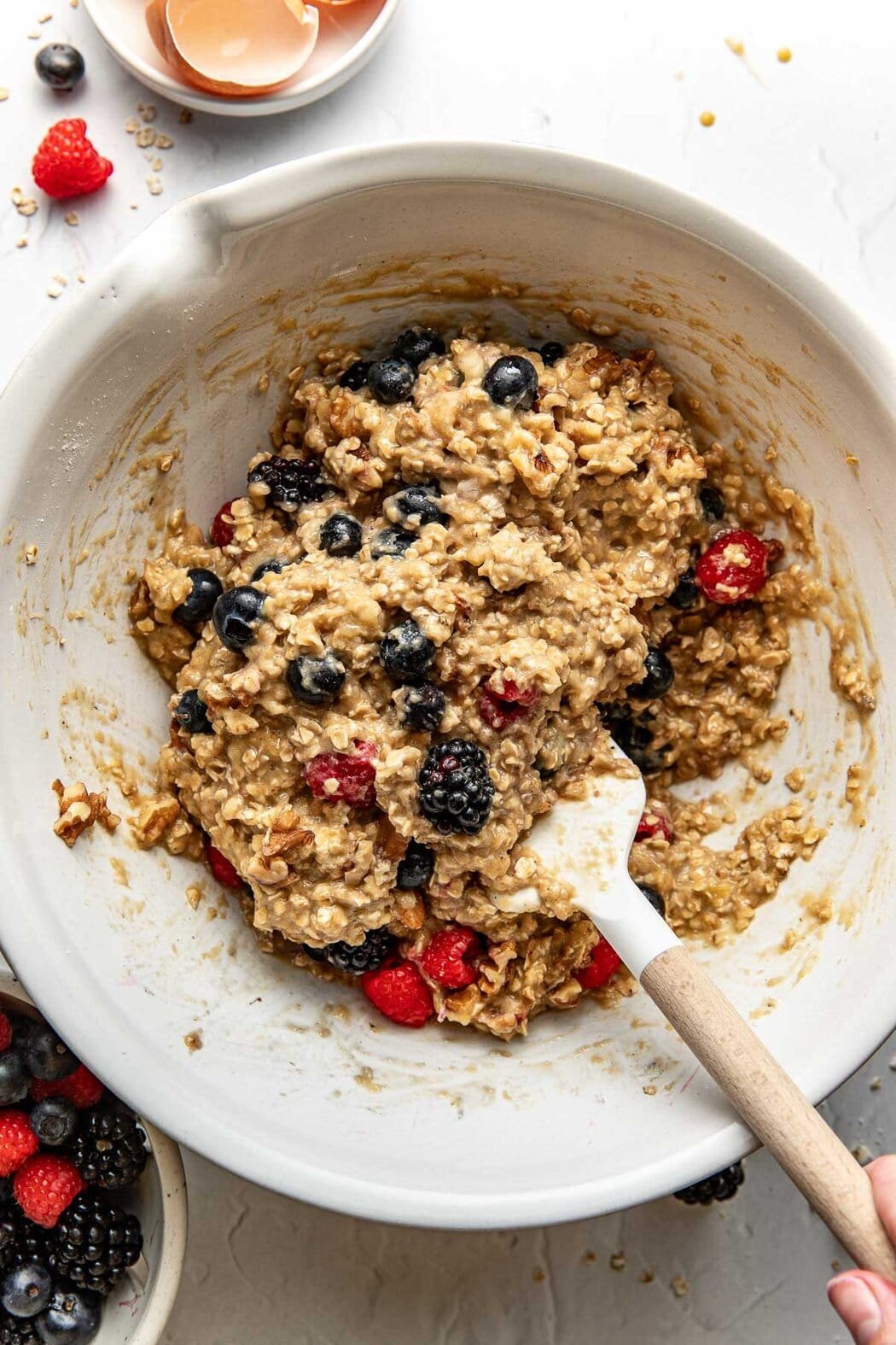 Overhead view of a white bowl of Mixed Berry Baked Oatmeal Muffin batter with a spatula on the side. 