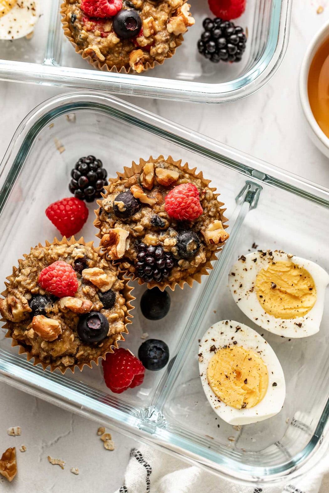 Overhead view of a glass meal prep container filled with two Mixed Berry Baked Oatmeal Muffins and a hard-boiled egg cut in half with berries on the side. 