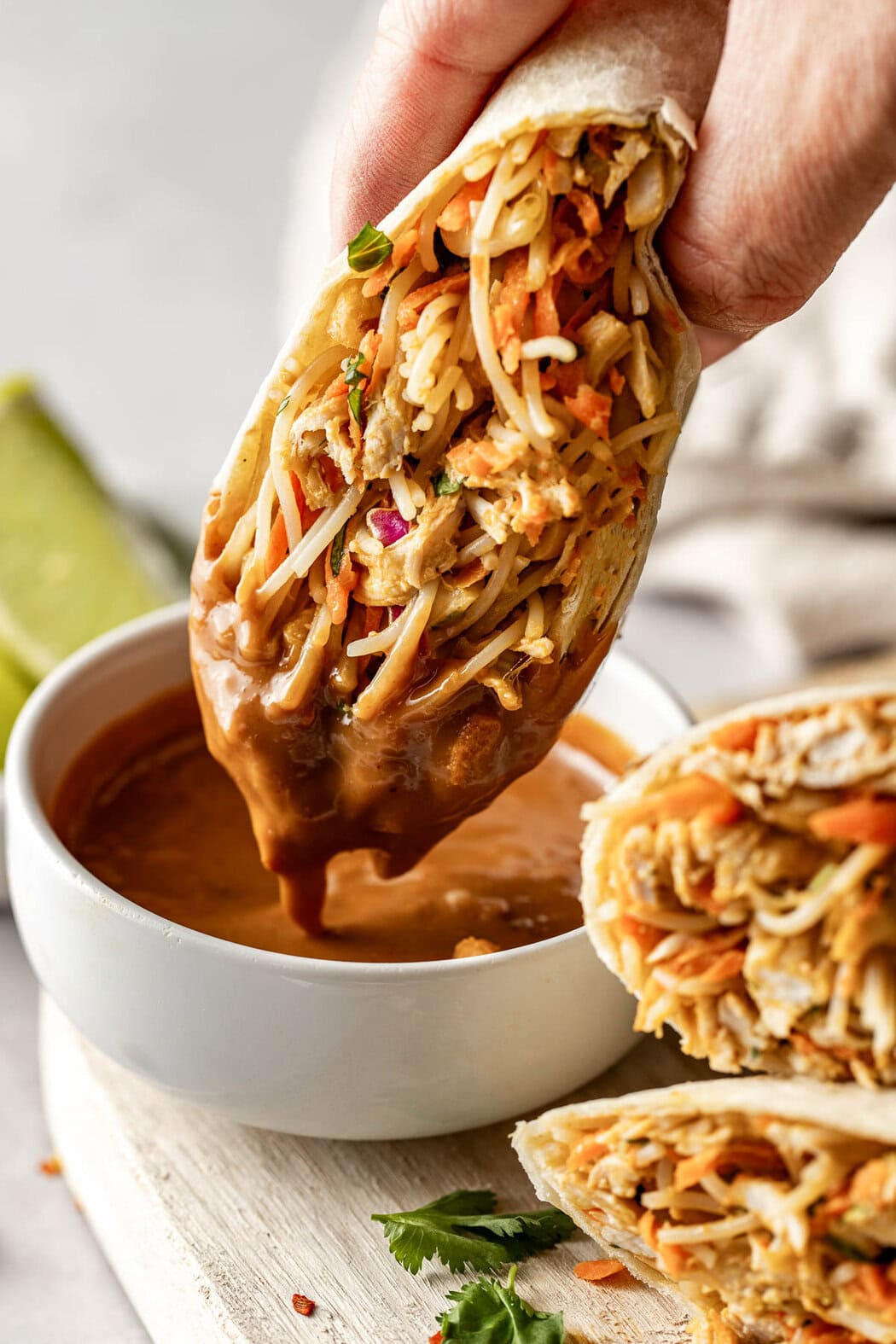 Close up view of a hand holding a Peanut Chicken Wrap and dipping it into a bowl of peanut sauce. 