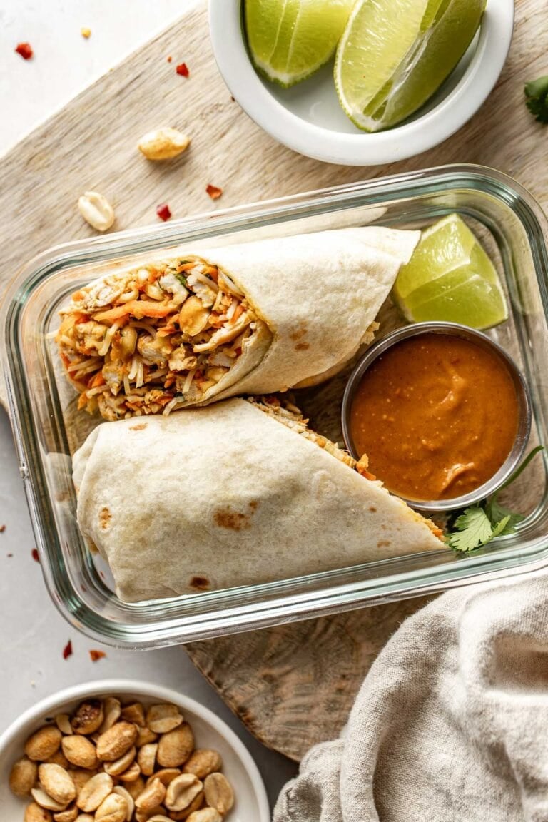 Overhead view of a glass meal prep container filled with a peanut chicken wrap cut in half with a small container of peanut sauce on the side.