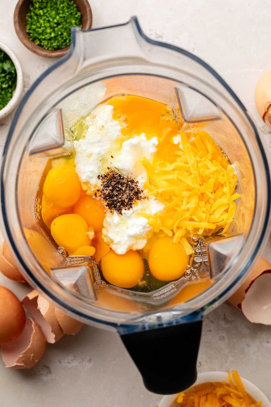 Overhead view of a blender filled with eggs, cottage cheese, shredded cheese, and black pepper. 
