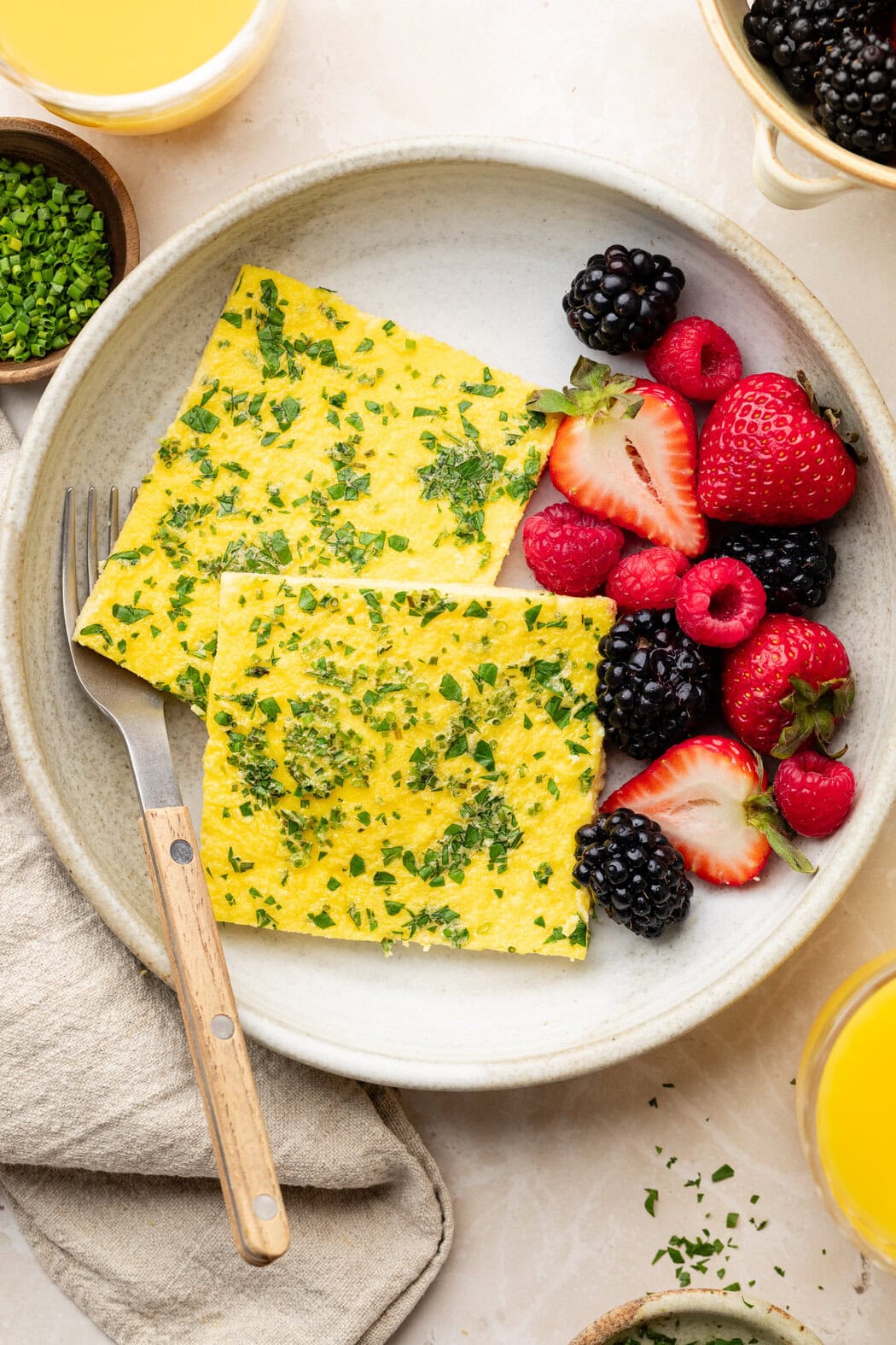 Overhead view of a white ceramic plate filled with 2 squares of sheet pan eggs topped with fresh herbs and colorful fruit on the side.