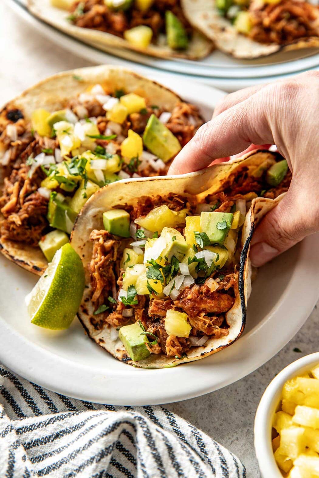 Close up view of a hand grabbing a tortilla filled with fresh Tacos Al Pastor and lime wedges on the side. 