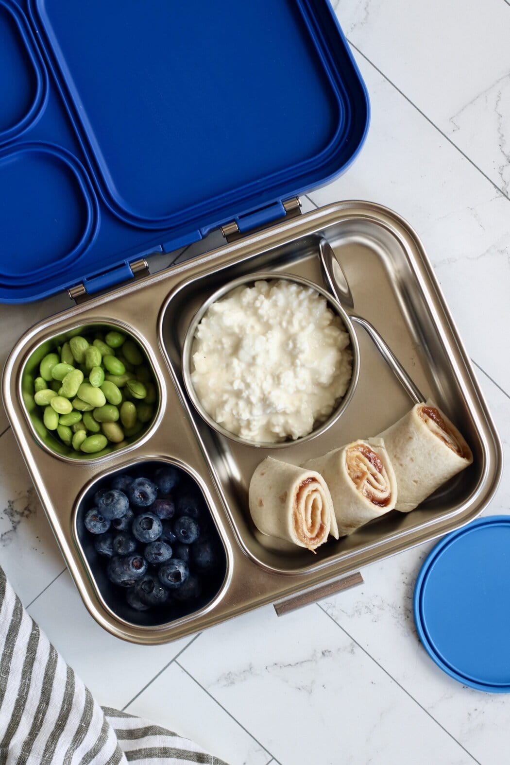Overhead view of an open lunch box filled with edamame, blueberries, cottage cheese, and peanut butter and jelly roll ups. 