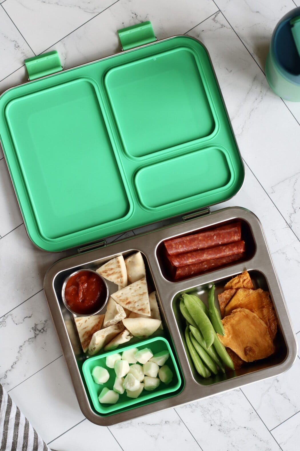 Overhead view of a lunchbox filled with pita pieces, mozzarella pearls, pepperoni, snap peas, and dried mango pieces. 