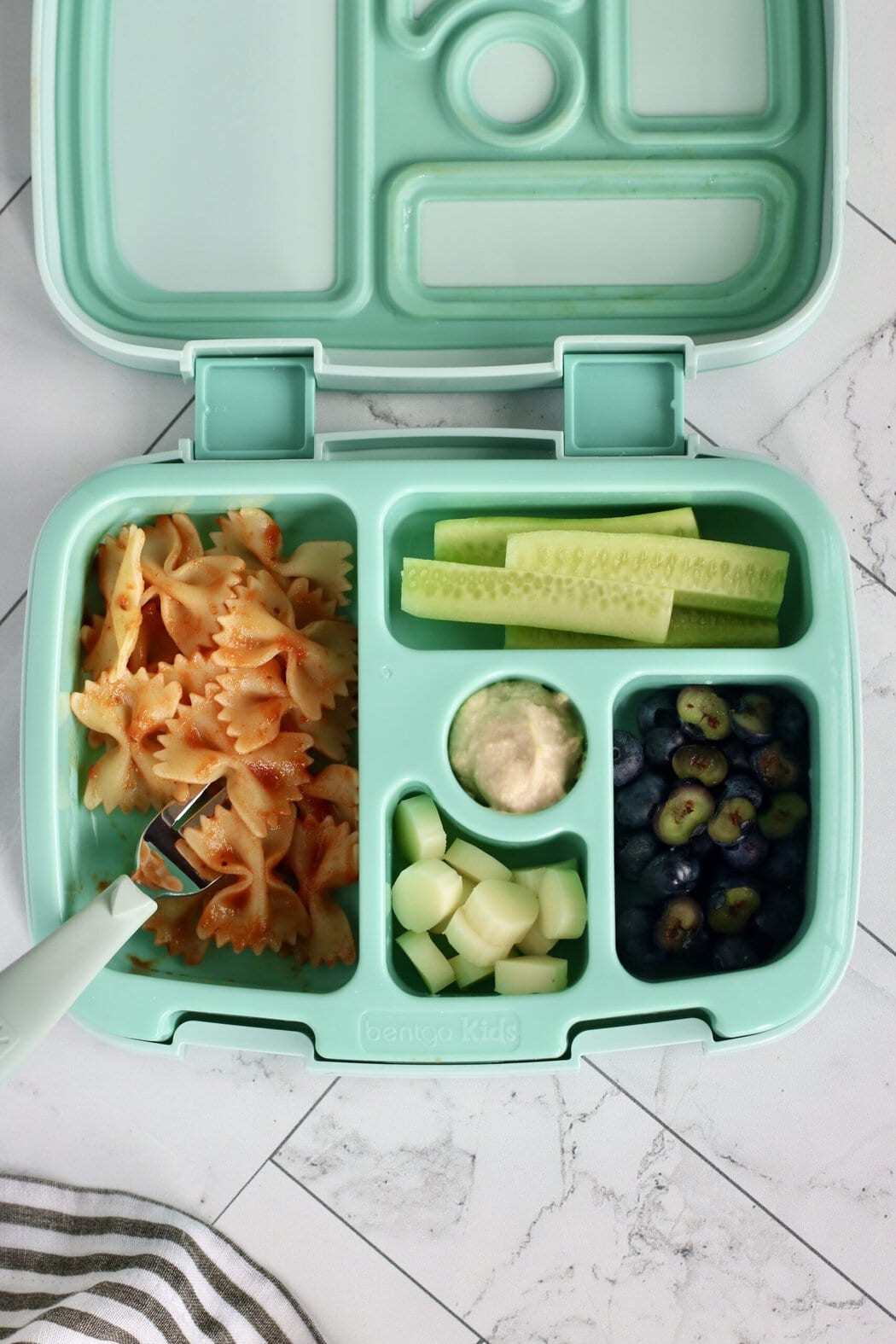Overhead view of an open lunchbox filled with bow tie pasta, cucumber sticks, blueberries, and mozzarella pieces. 