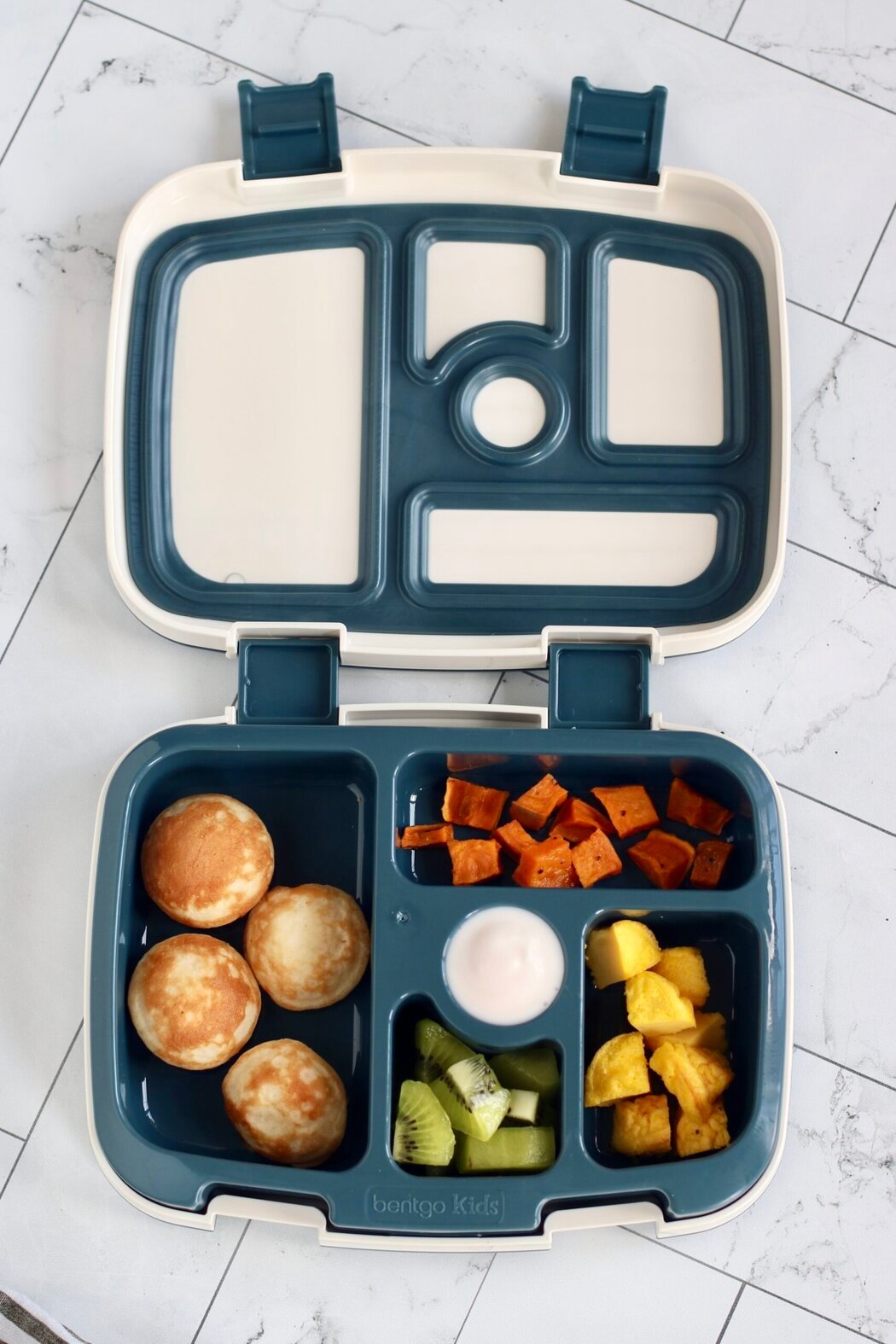 An open lunchbox filled with mini protein pancakes, cut up eggs bites, roasted sweet potato pieces, and kiwi pieces. 
