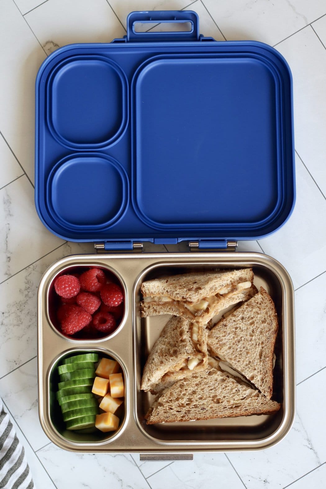 Overhead view of an open lunchbox filled with red raspberries, cucumber slices, cheese cubes, and a peanut butter and banana sandwich cut into four pieces. 