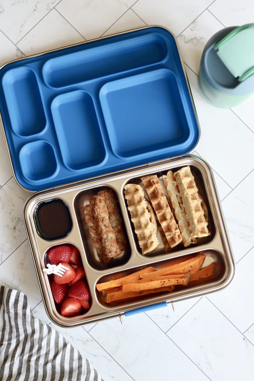 Overhead view of an open stainless steel lunch box filled with waffle sticks, sausage links, strawberries, and roasted sweet potato fries. 