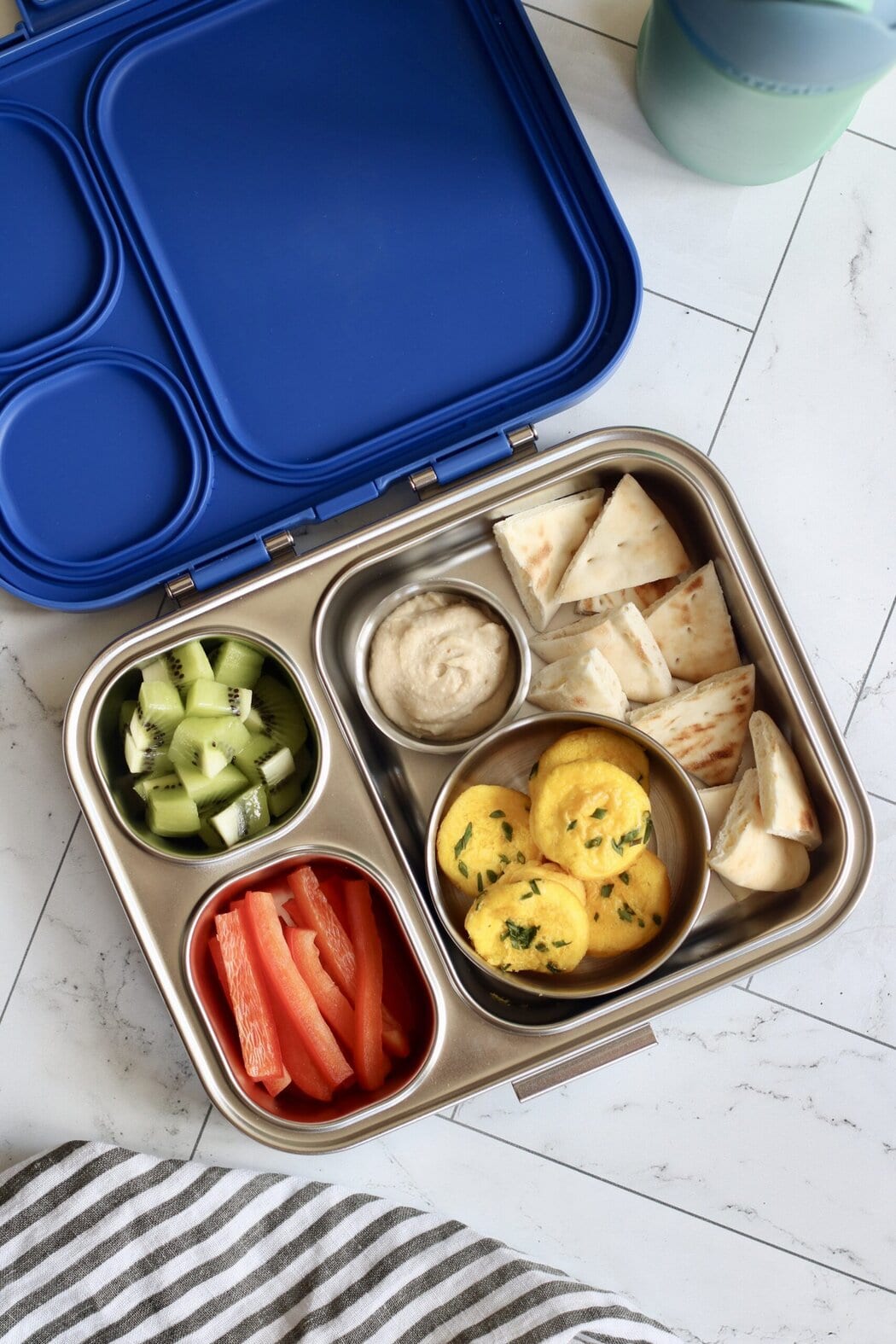 Overhead view of an open lunchbox filled with kiwi fruit, red pepper strips, pita and hummus, and cottage cheese egg bites topped with fresh chives. 