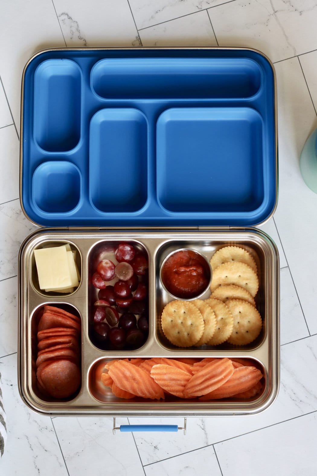 Overhead view of an open stainless steel lunchbox filled with sliced grapes, carrot chips, crackers, marinara sauce, sliced cheese, and pepperoni. 
