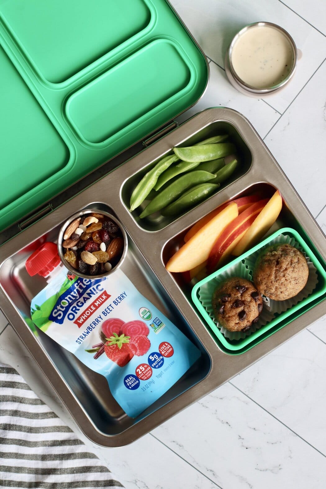 Overhead view of an open lunchbox filled with a yogurt pouch, trail mix, snap peas, peach slices, and two mini muffins. 
