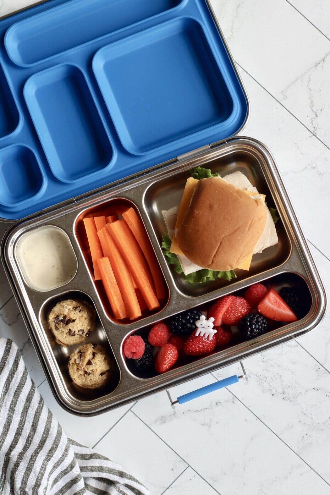 Overhead view of an open lunchbox filled with chickpea cookie dough bites, carrot sticks, berries, and a slider sandwich. 