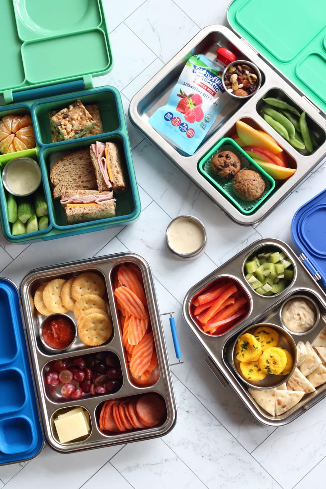 Overhead view of four different lunchboxes open and laying on a marble countertop, showing a variety of colorful healthy snack options. 