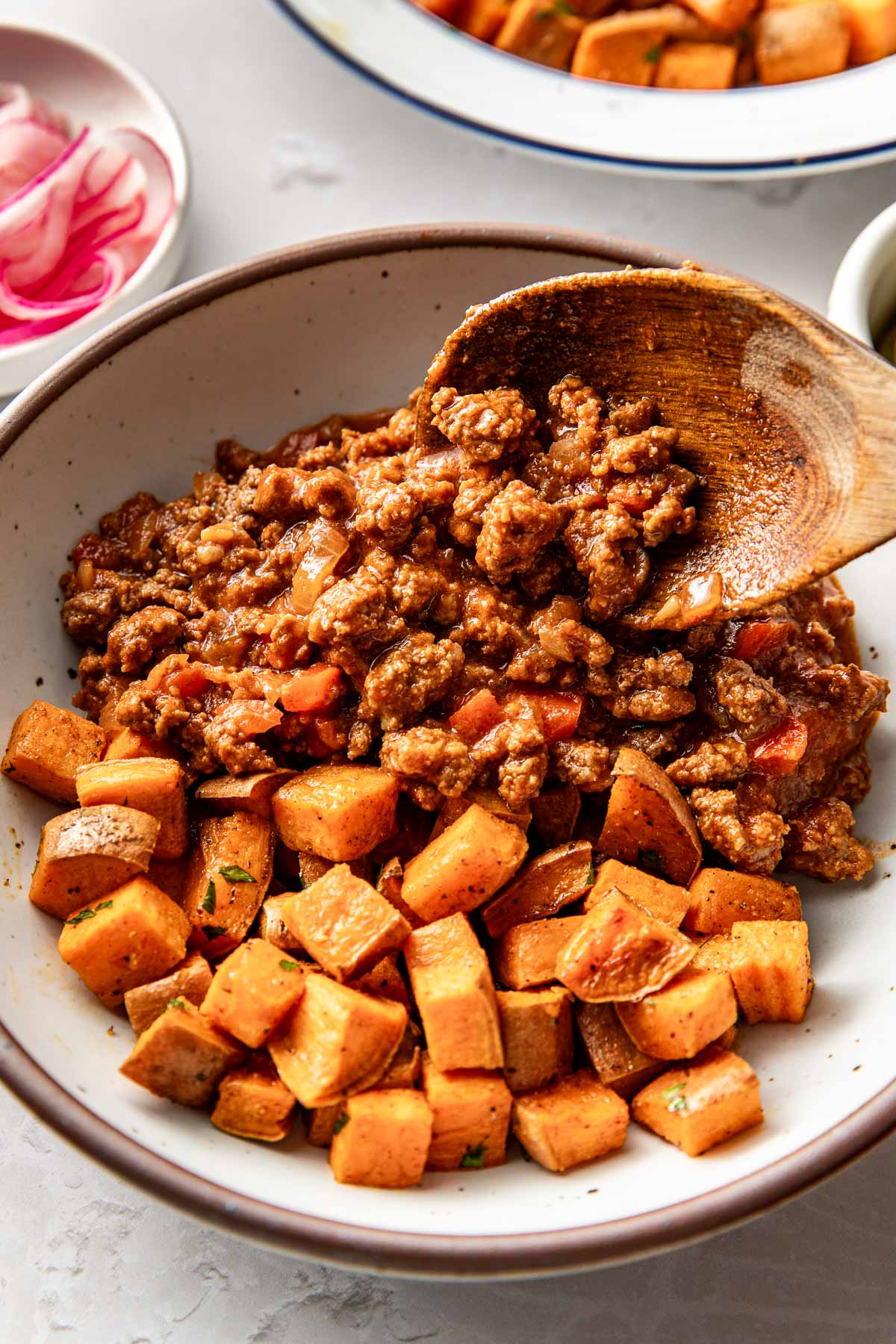 Close up view of a bowl of roasted sweet potatoes with a wooden spoon placing sloppy joe meat over the top. 