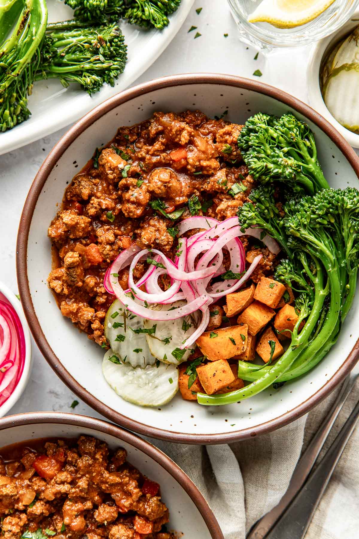 Close up view of a plate of sloppy joe meat with roasted sweet potatoes, broccolini, pickled red onions, and crisp pickle chips on the side. 
