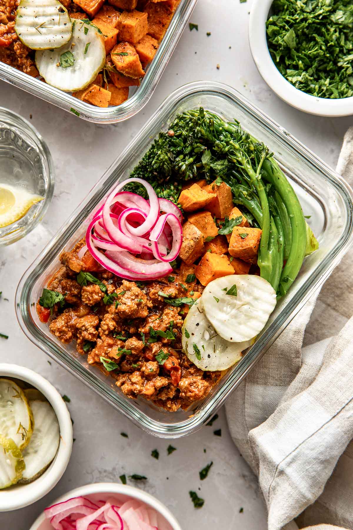 Overhead view of a glass meal prep container filled with Sloppy Joe Meat, roasted sweet potatoes, broccolini, pickled red onions, and crisp dill pickle chips topped with fresh herbs. 