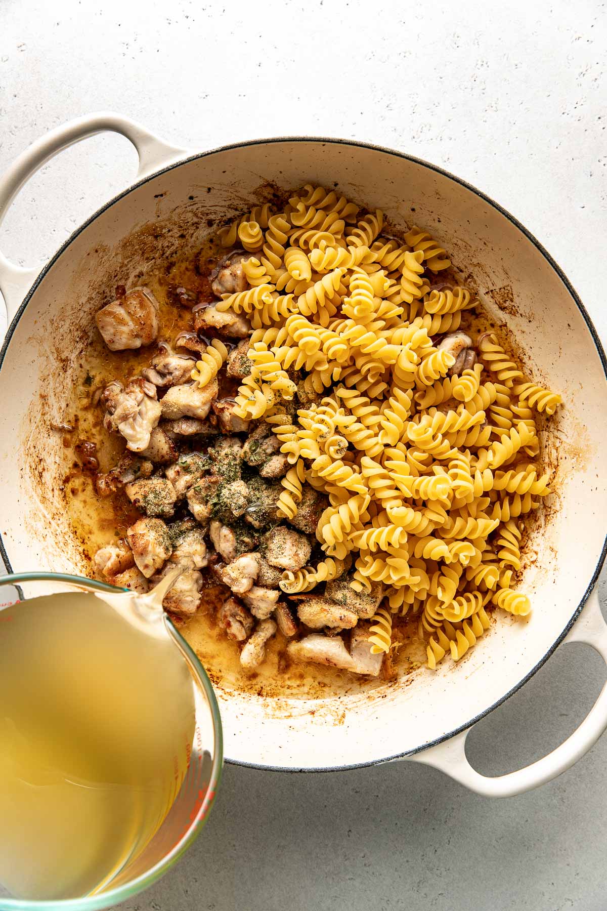 Overhead view of a skillet filled with cooked diced chicken and pasta with a hand pouring broth over it. 