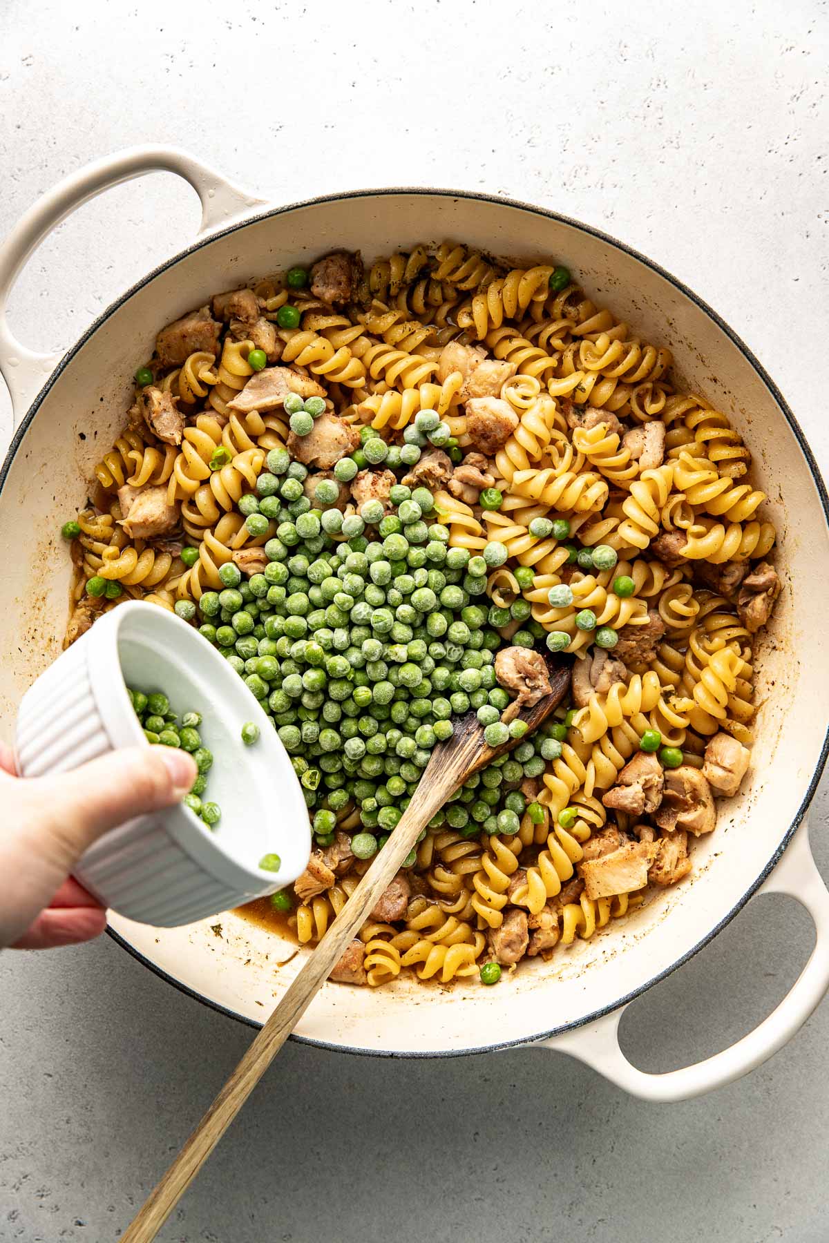Overhead view of a skillet filled with cooked pasta and diced chicken with a hand pouring a bowl of fresh peas on the top. 