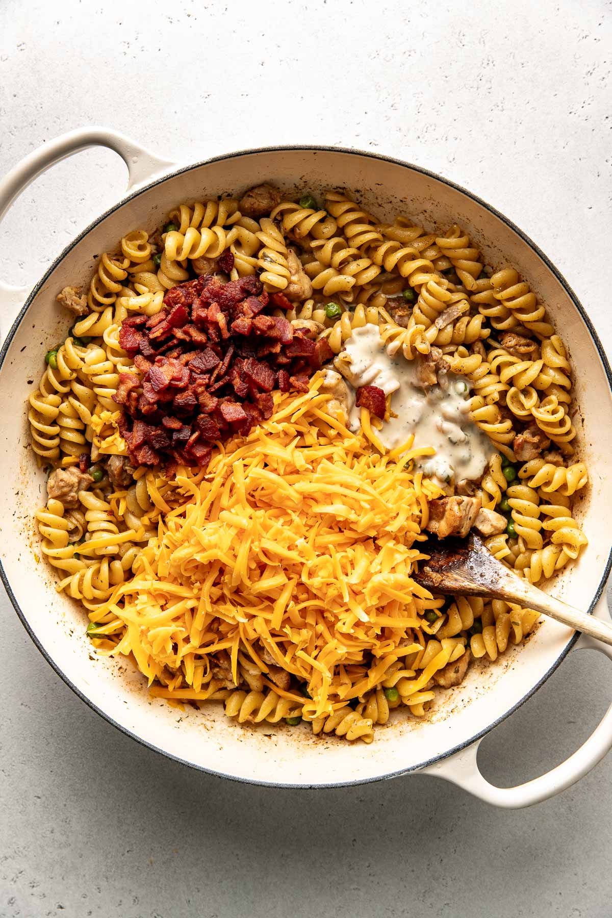 Overhead view of a skillet filled with cooked pasta, diced chicken, crumbles of bacon and topped with shredded cheddar cheese ready for stirring. 