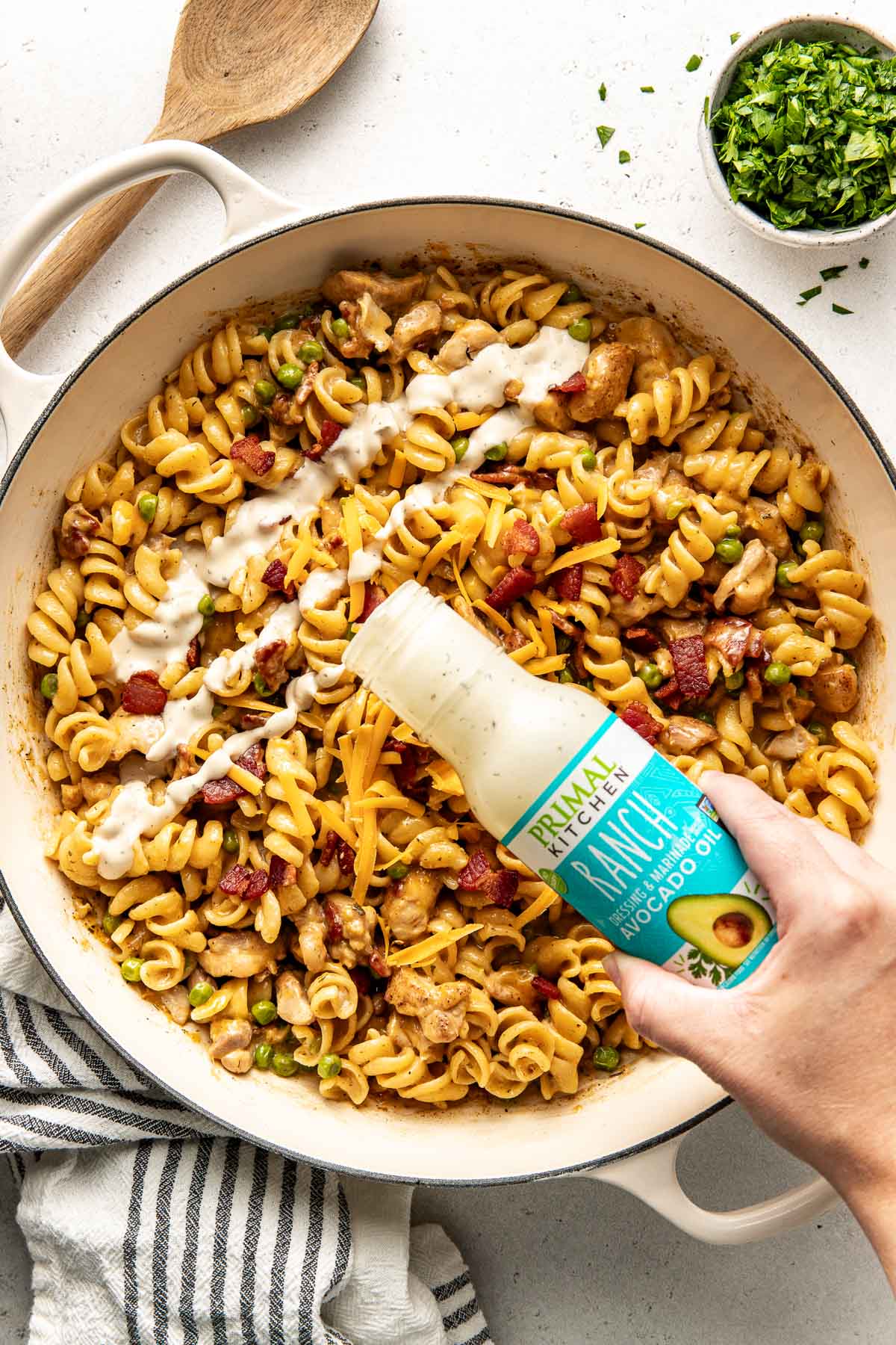 Close up view of a skillet filled with One-Pot Chicken Bacon Ranch Pasta with a hand pouring primal kitchen ranch dressing on the top.